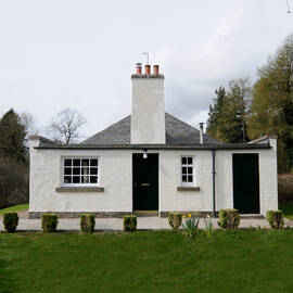A small white cottage with a tall stone chimney and black painted doors. The cottage is surrounded by a grassy lawn and little box shrubs.