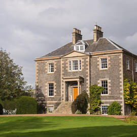 A three-storey grand Georgian house stands at the head of a large garden. A grass lawn lies in front, framed by tall trees.
