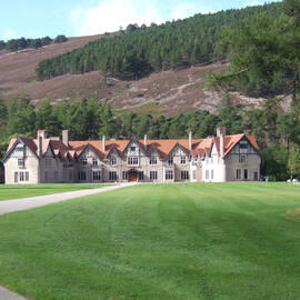 A view of Mar Lodge, with its distinctive red tiled roof. A long driveway sweeps across a grassy field in front. Heather- and pine-covered hillside rises behind.