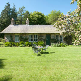 A small Edwardian cottage with a large green lawn in front. Blossoming trees frame the image. A garden table and chairs are set up in the middle of the lawn.