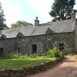 A row of stone cottages with a gravelled driveway leading up to it. A green lawn bordered by a low stone wall lies in front.