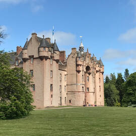 A view of the exterior of Fyvie Castle, looking towards one of the corner towers from across the smooth grass lawns. Tall trees stand either side of the castle.
