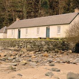A long white single-storey cottage stands just above a sandy beach, with pebbles across it. A steep hillside rises behind the cottage.