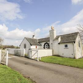 A single-storey, white walled cottage stands beside a white wooden gate at the entrance to a driveway. Grass runs alongside the driveway.