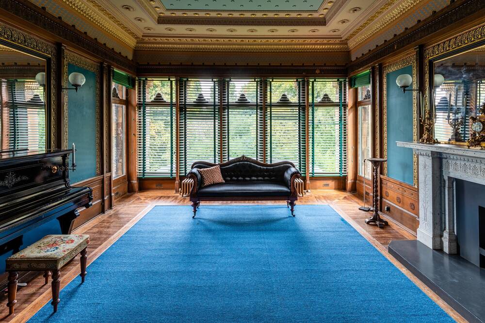 A view looking along the length of a grand drawing room to the large bay windows at the far end. The walls are wood panelled with blue and gold decoration. A large blue rug covers the floor. A leather sofa with carved wooden legs stands just inside the bay.