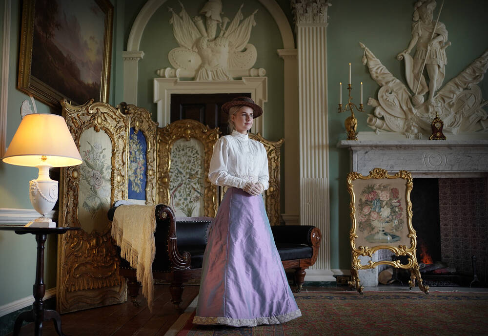 A costumed guide dressed as a Victorian lady in a high-collared, long-sleeved white and purple dress and a hat is stood in the Saloon at House of Dun. Behind her is an ornate three-piece screen and fireplace. The walls are adorned with intricate plasterwork designs.
