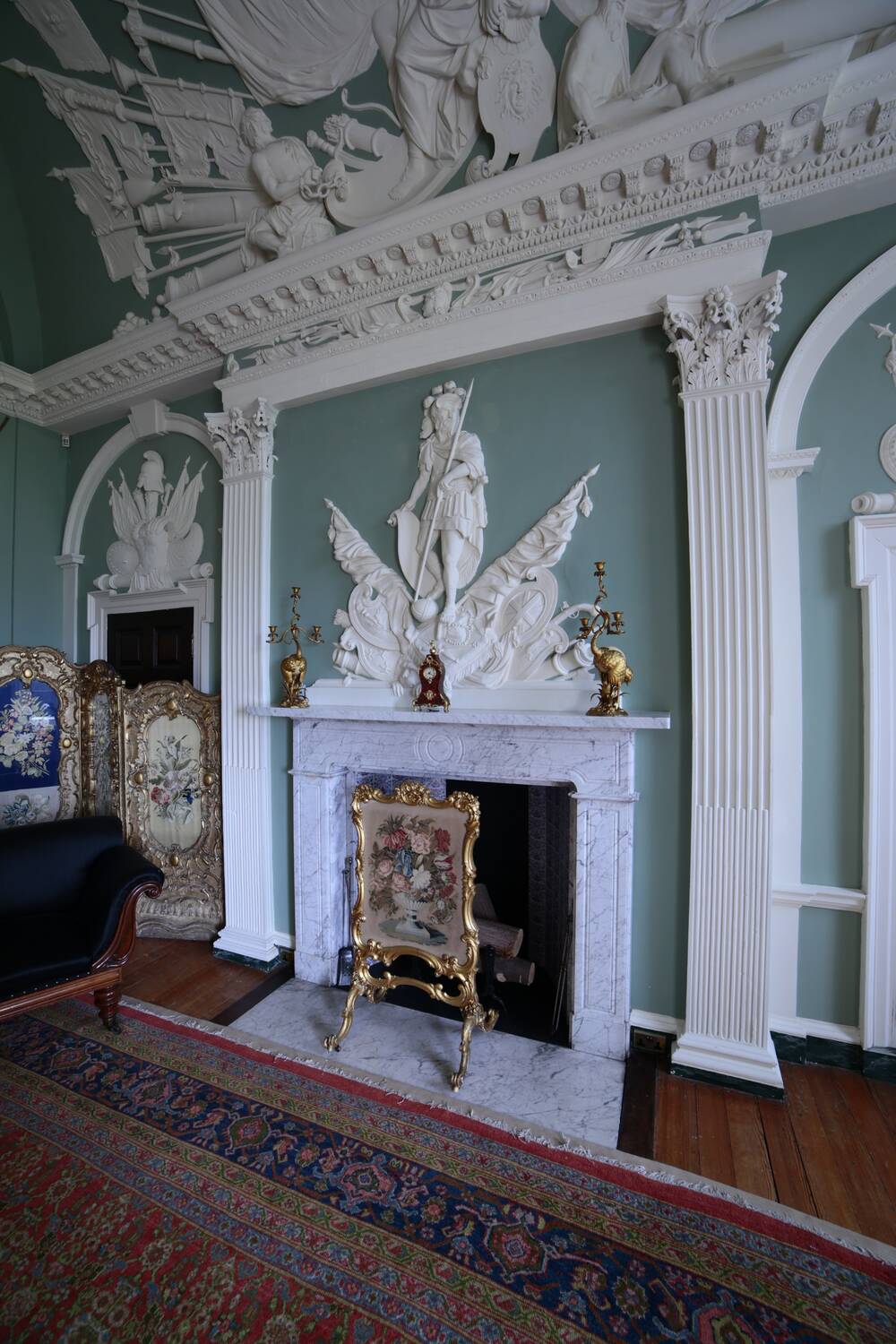 A view of elaborately carved plasterwork on teal-blue walls in a grand drawing room in a country house. The panel above the fireplace features Mars with one foot resting upon a round crown. The plaster design continues onto the ceiling, where more classical scenes can be seen. An embroidered fireguard stands on the fireplace.