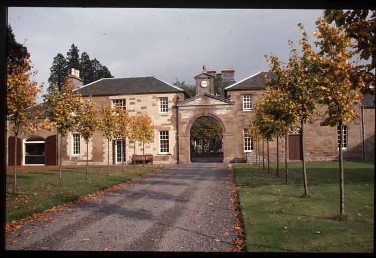 A view of the stable block at House of Dun, with a gravel path leading towards a stone arch with a clock above.
