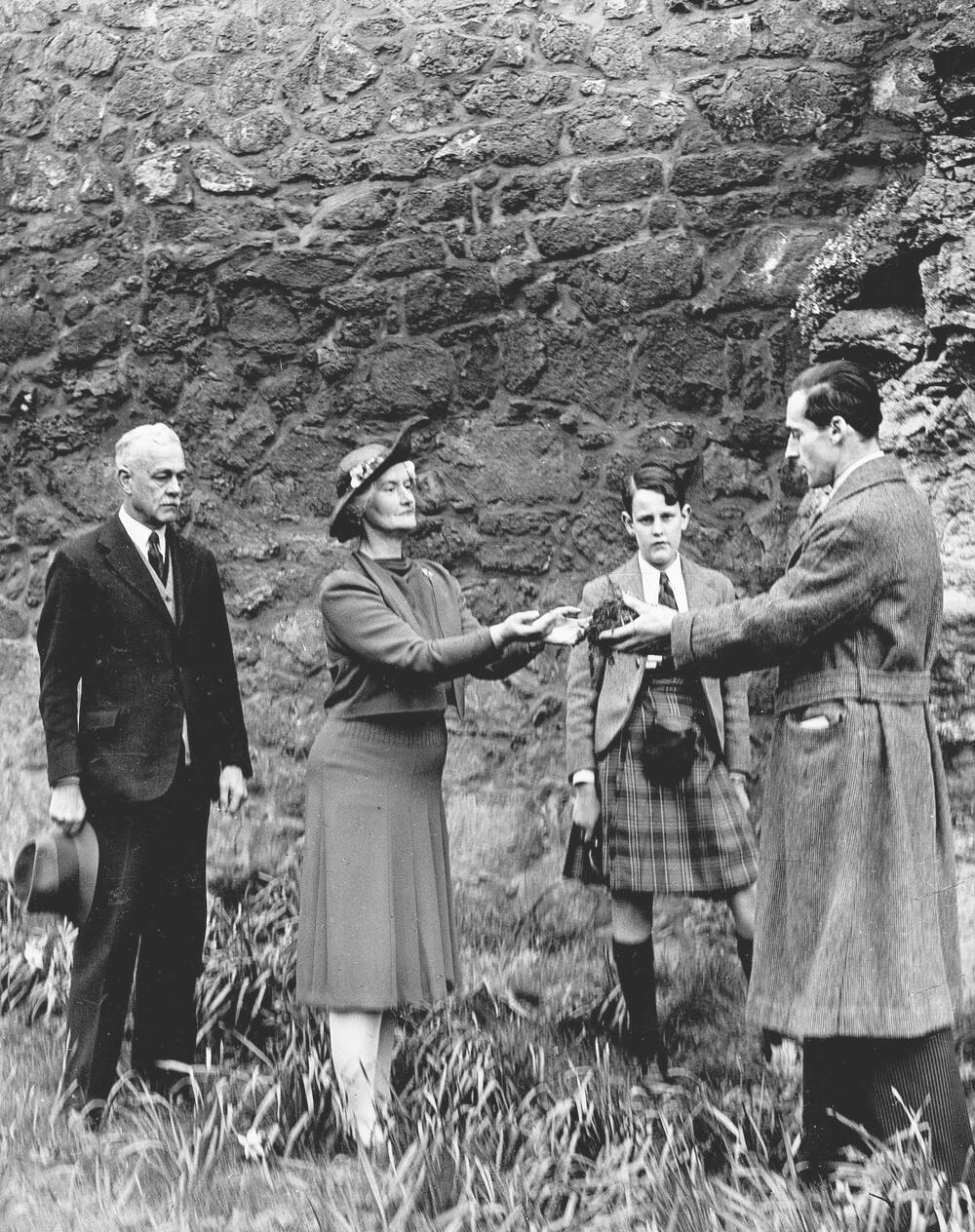 A black and white photo of four people standing beside an old, very tall stone wall. A smartly dressed couple hand over a large key to a man wearing a long wool coat. In between them stands a boy wearing a kilt, who watches the ceremony.