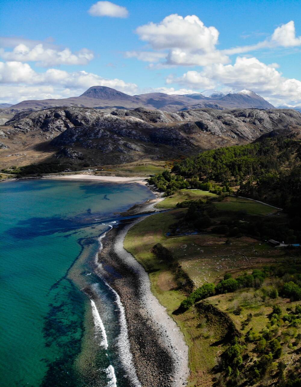 An aerial view of Inverewe Garden focusing on the shoreline by the loch. The water is turquoise and clear. Mountains can be seen in the distance.