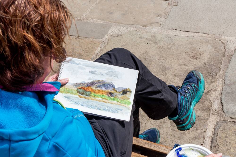 A woman sits on a wooden bench with a sketch pad lying across her knee. On it is a watercolour of a loch with mountains beyond. The photo is taken looking over the woman's shoulder.