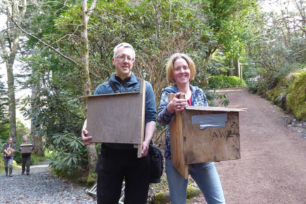 A man and a woman stand beside each other at a junction of several garden paths. Both hold large wooden feeder boxes in their arms. Behind them on the path are two more people, also both carrying large wooden boxes.