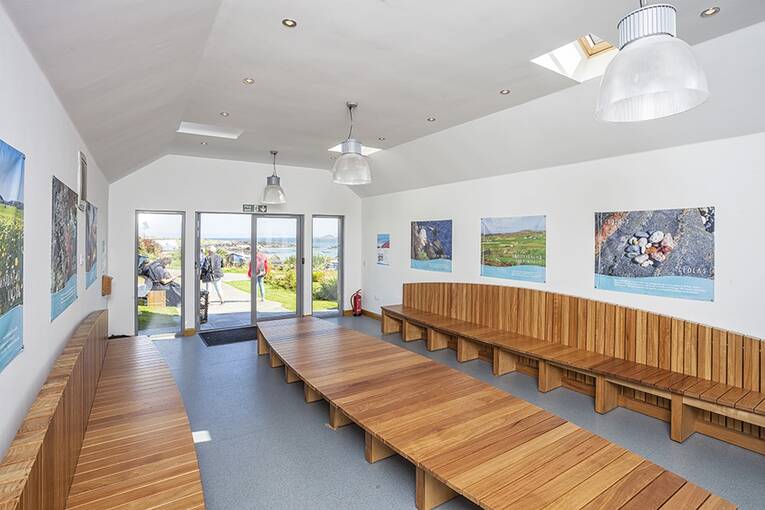 A view inside the Iona Ferry Shelter, showing long wooden benches running the length of the room. Posters and panels brighten up the white walls.