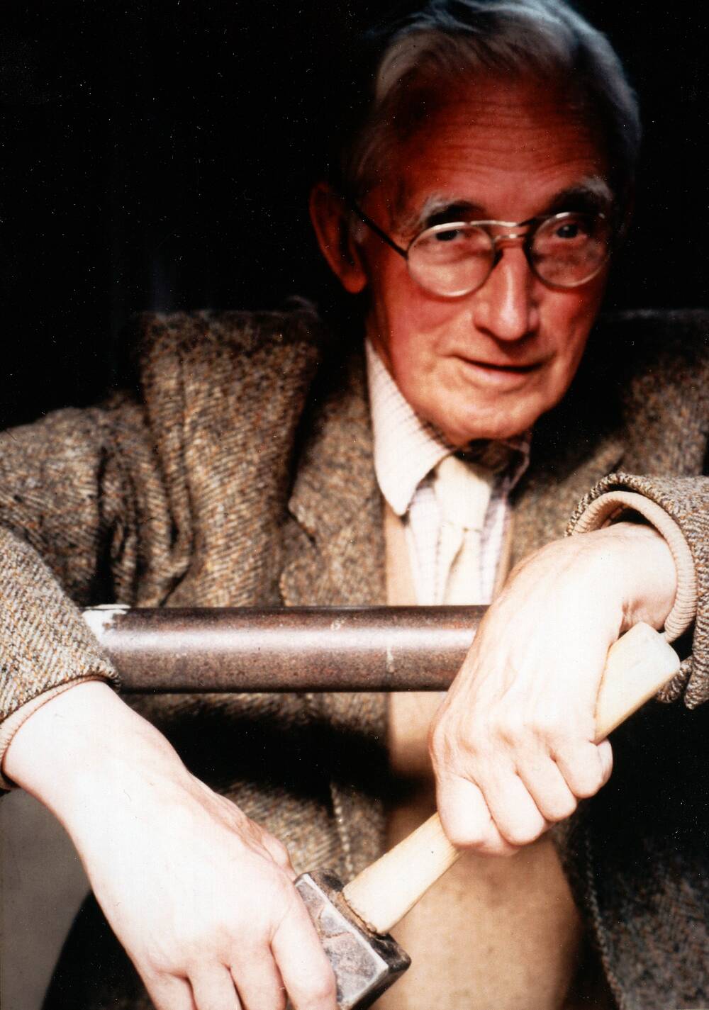 An old colour photo of Hew Lorimer, who stands and rests his arms over a metal railing. He is holding a small mallet in both hands. He wears glasses and a brown tweed jacket. He has a wry smile.