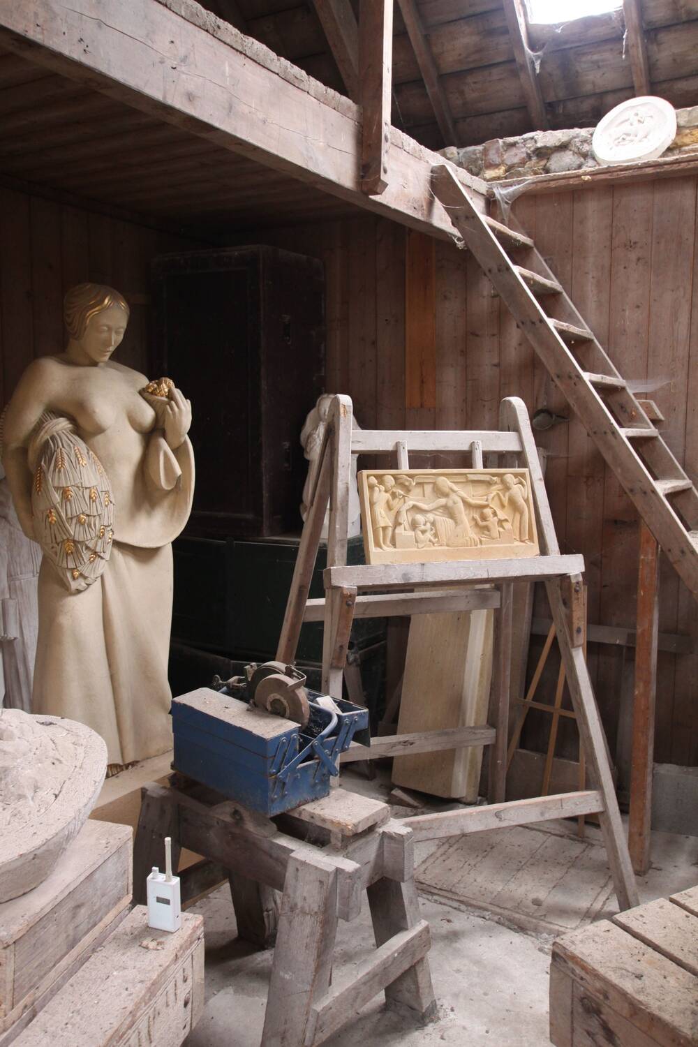 A view of an artist's sculpture studio, with a ladder leading up to a wooden loft area above. A tall statue of a woman with golden hair holding a sheaf of wheat stands in the corner. In the foreground are wooden steps, easels and a blue toolbox.