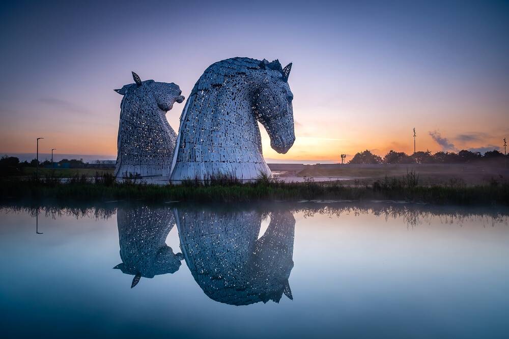 Two very large metal sculptures of horse heads loom into the dusk sky. They are reflected almost perfectly in the still water in the foreground.