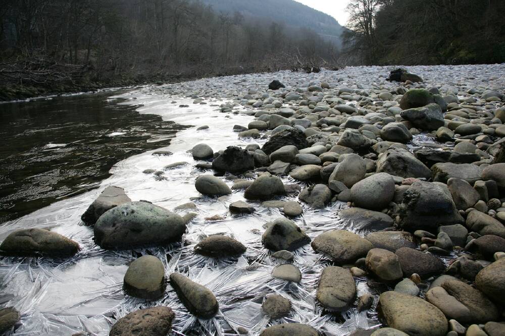 A view taken from almost ground level looking out across a wide river, which is almost completely frozen over. Large stones are at the edges in the foreground, with cracked patterns of ice linking them together.