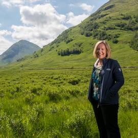 A smiling woman stands on a grassy hillside, with dramatic mountains behind her. She has shoulder-length fair hair and wears a National Trust for Scotland outdoor jacket.