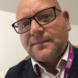 A close-up view of a man looking down into the camera. He has no hair and wears black-framed glasses. A purple National Trust for Scotland lanyard hangs on top of a navy jacket and open-necked shirt.