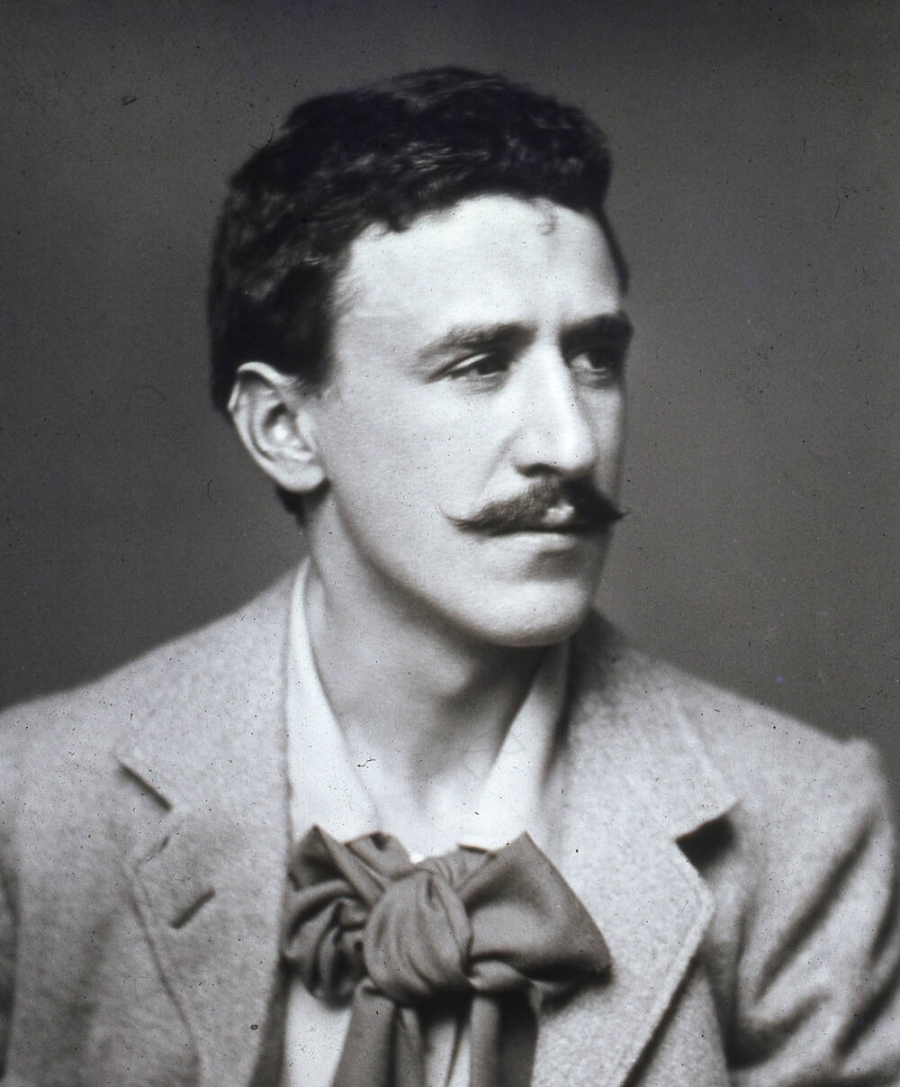 A black and white photograph of Charles Rennie Mackintosh​​, wearing a large cravat and showing his distinctive moustache.