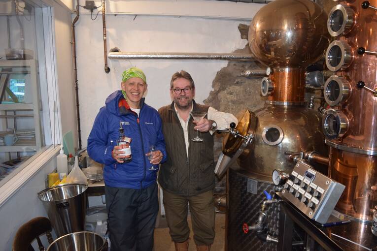 A man and a woman stand side by side, smiling at the camera. One holds a bottle of gin and the other a glass. They are in a distillery beside gin-making equipment.