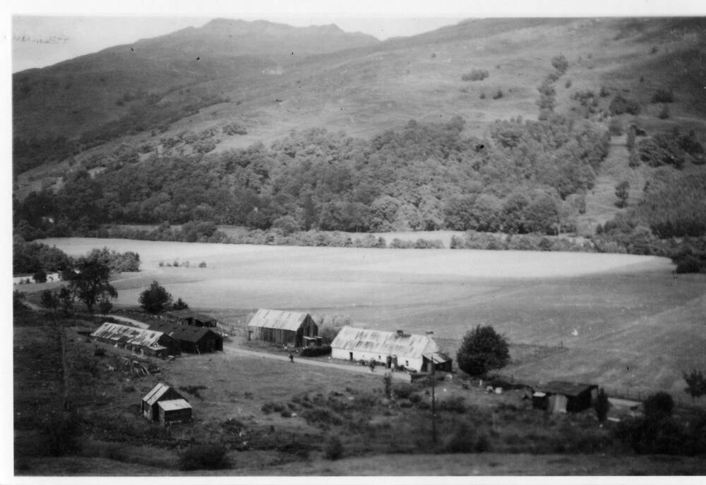 A black and white photograph of Moirlanich Longhouse in the 1930s, showing a lean-to dairy at the side. A large barn stands to the other side. On the opposite side of the road is a collection of other farm buildings. There is a large flat field behind the cottage, and behind it a wooded hillside.