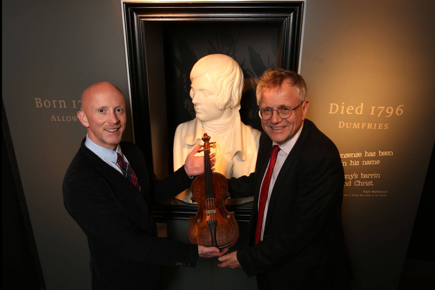 Two men hold the Burns violin between them, standing in front of a bust of Burns.