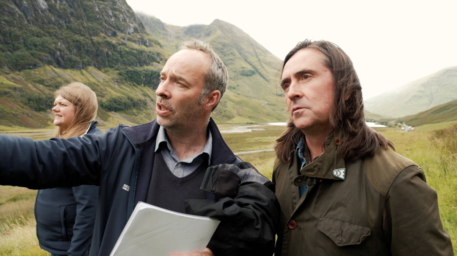 Neil Oliver stands beside Derek Alexander, the Trust's Head of Archaeology, as they both look down the glen at Glencoe. Derek holds some papers under his arm and is pointing ahead of him.