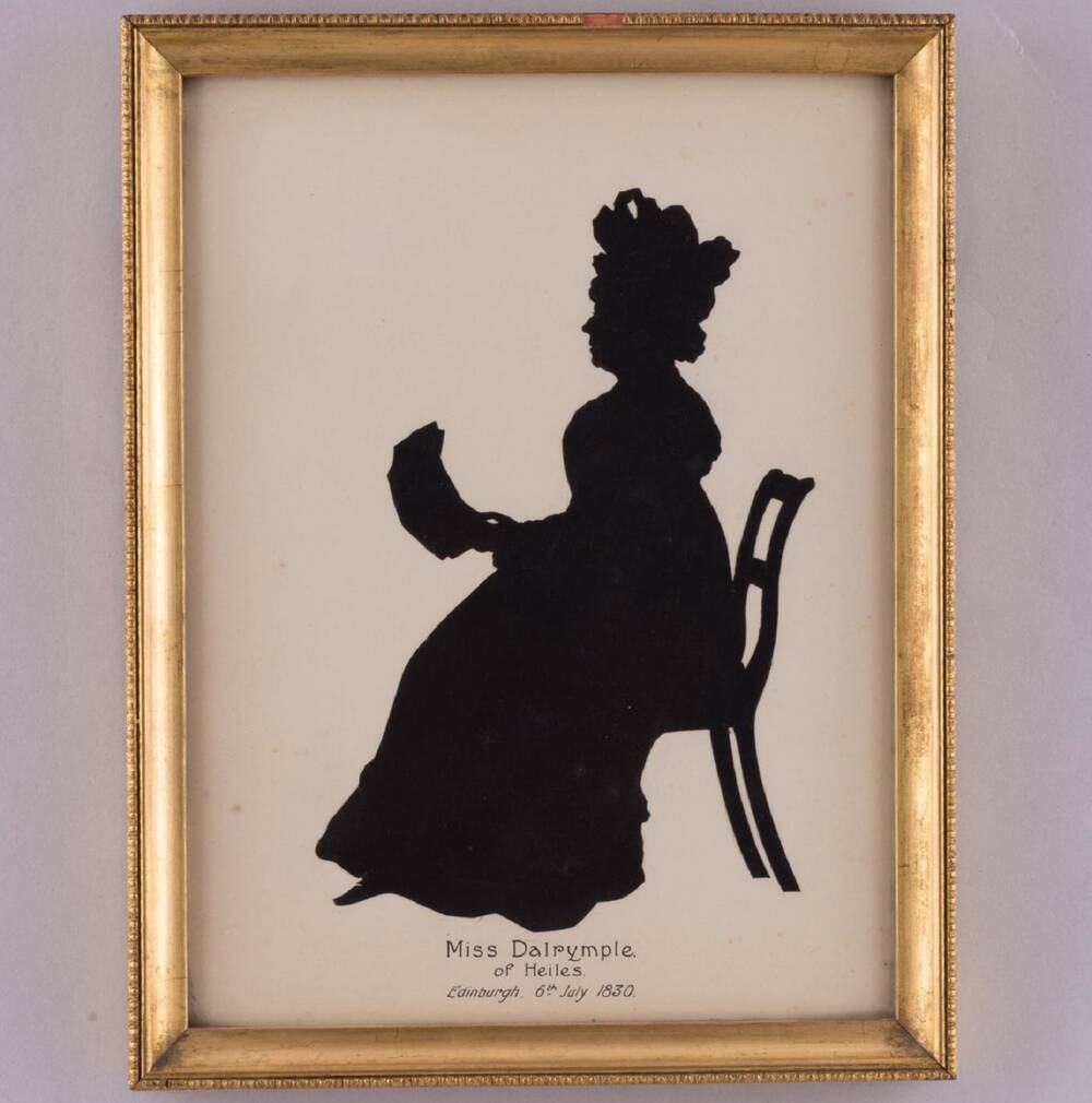 A framed silhouette portrait of an older lady sitting in profile on a chair. She holds something like a fan on her lap. She is wearing an elaborate hat.