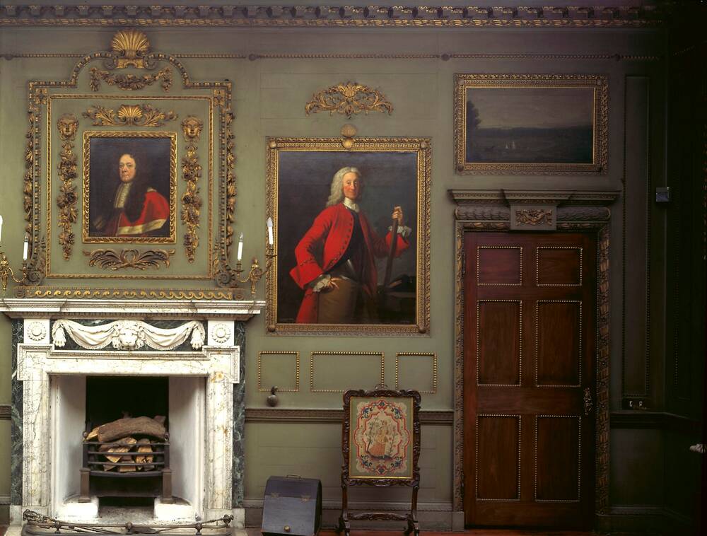 A view of a green painted wall in a very grand house. The wall has a number of gold-framed portraits hanging on it, above a white marble fireplace. A tapestry fireguard stands beside the fireplace.