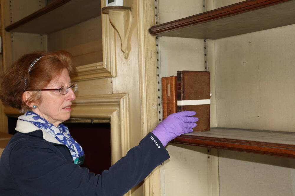 A lady wearing purple conservation gloves carefully places old books onto a shelf.