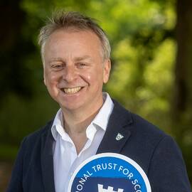 A photo of a smiling man, wearing a navy jacket and white shirt. He is holding an omega-shaped sign, with the National Trust for Scotland logo and the word Love on it. Green trees are in the background.