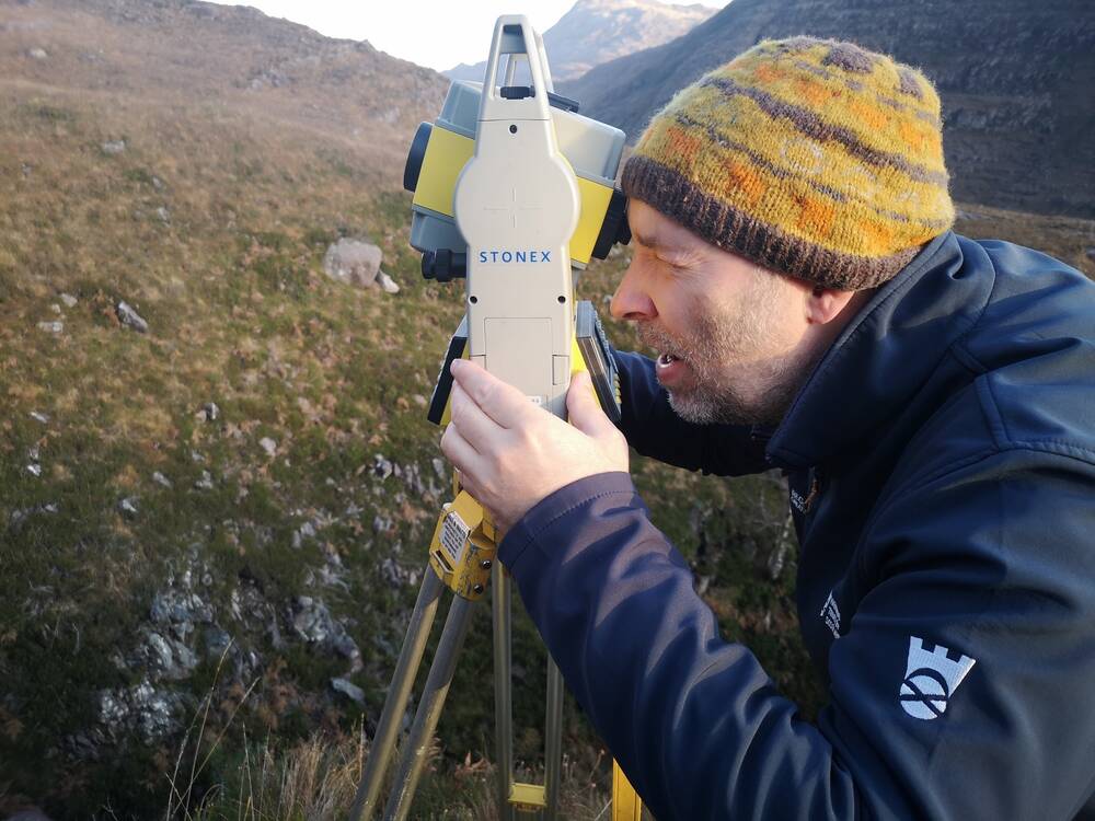 A man wearing a woolly hat looks through a camera-like device on a tripod. This is set up in a moor-type area with mountains visible in the background.