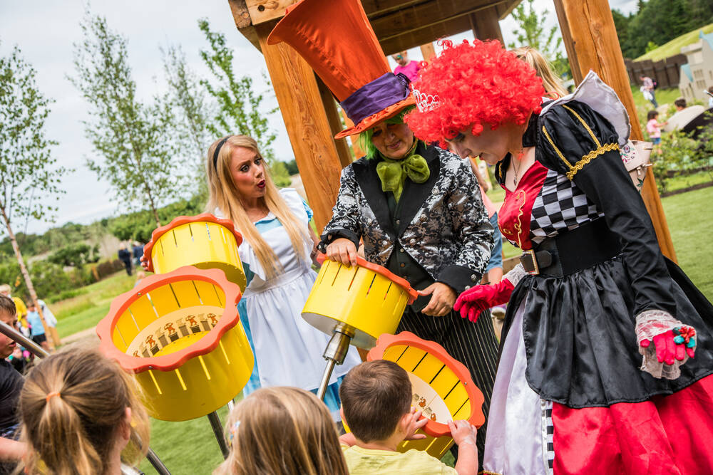 Three adults dressed as characters from Alice in Wonderland talk to a group of children in a play area. They are standing beside some giant yellow sculptures of daffodils.
