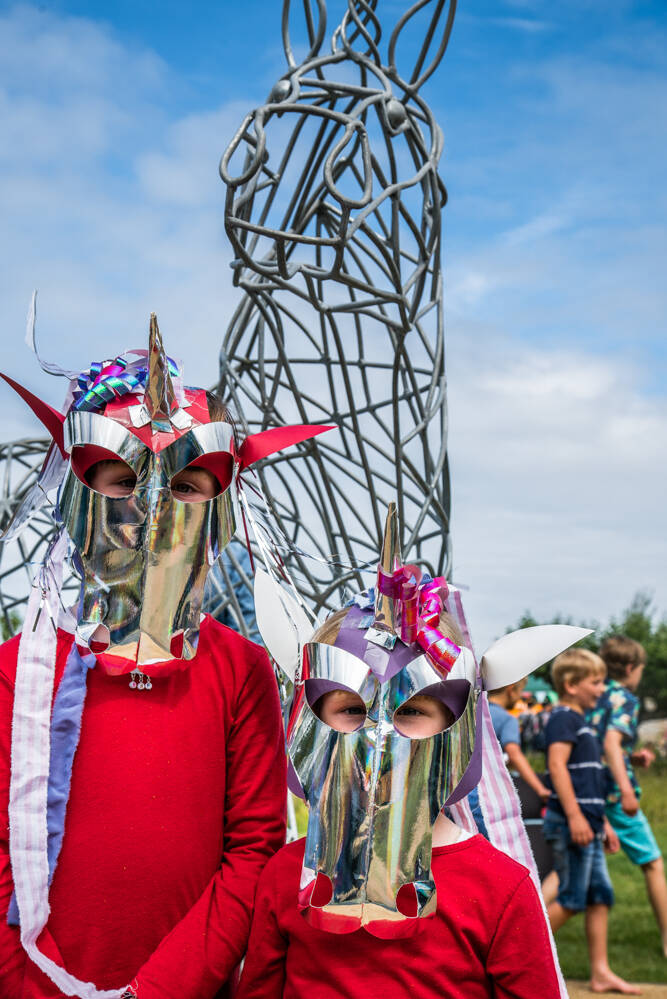 Two children wearing foil unicorn masks stand in front of a large metal sculpture of a unicorn in a playground. The masks also have colourful streamers tied to their unicorn horns.