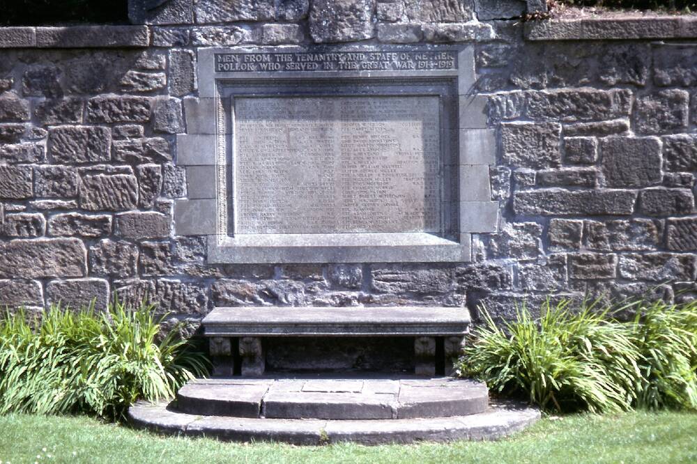 A large stone plaque, listing many names, is set into a stone brick wall. A stone bench stands just in front of the plaque. Decorative grasses grow either side.