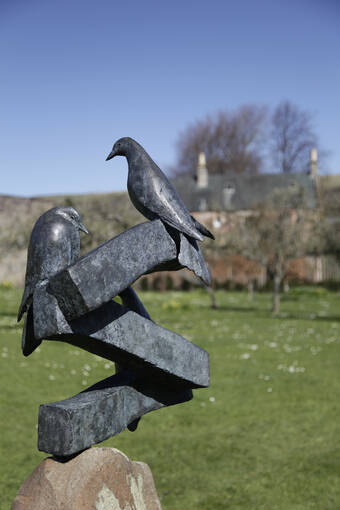 A metal sculpture of two doves is balanced on a rock pedestal, with a large garden lawn in the background.