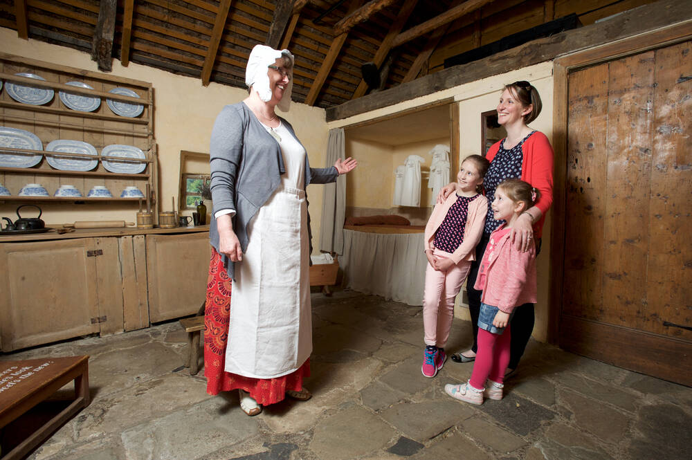 A mum and two girls stand in Burns Cottage looking at a costumed guide, dressed in a Georgian cap and apron..