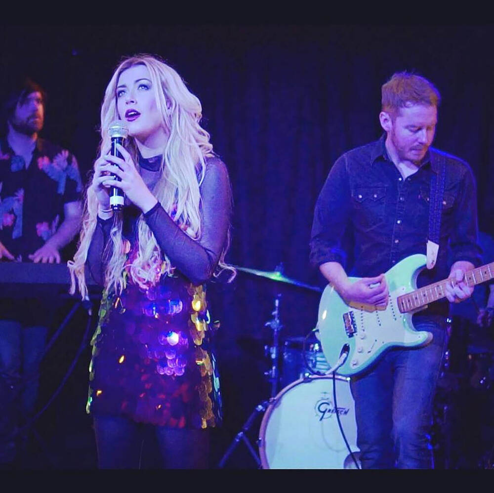 A young woman with long blonde hair stands on stage, holding the mike with both hands as she sings. Behind her a young man plays the electric guitar, and another young man plays the keyboard.