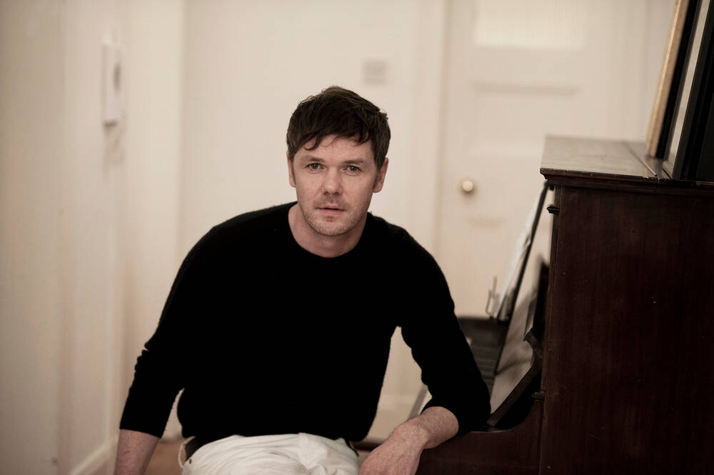 Roddy Woomble, the lead singer of Idlewild, sits at a piano. He has turned away from the keys to face the camera, and rests his left arm along the keys.