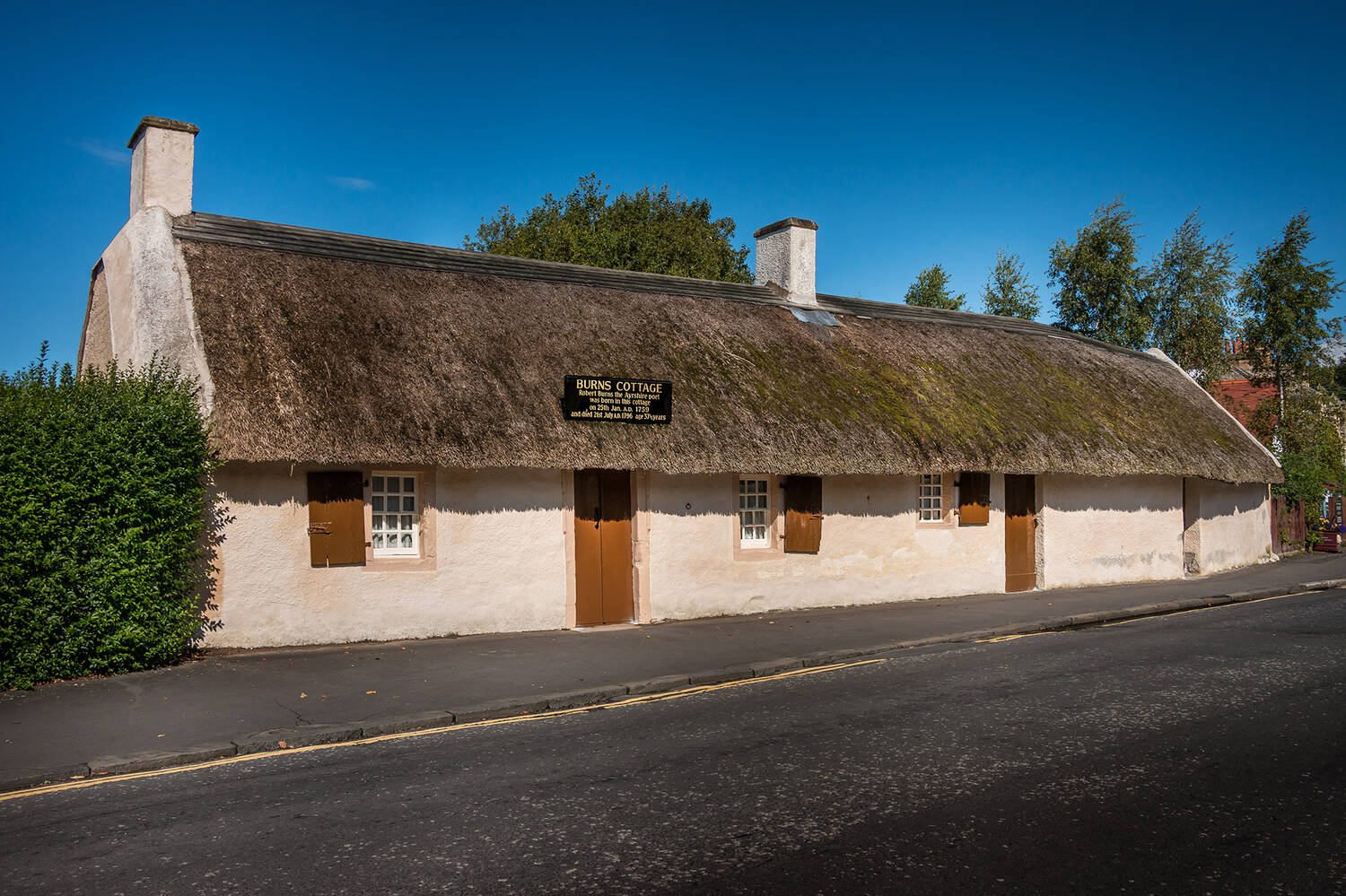 A view of the exterior of Burns Cottage, a single-storey long cottage with white stone walls and a thatched roof. A road passes directly in front of it. The sky is a bright blue behind.