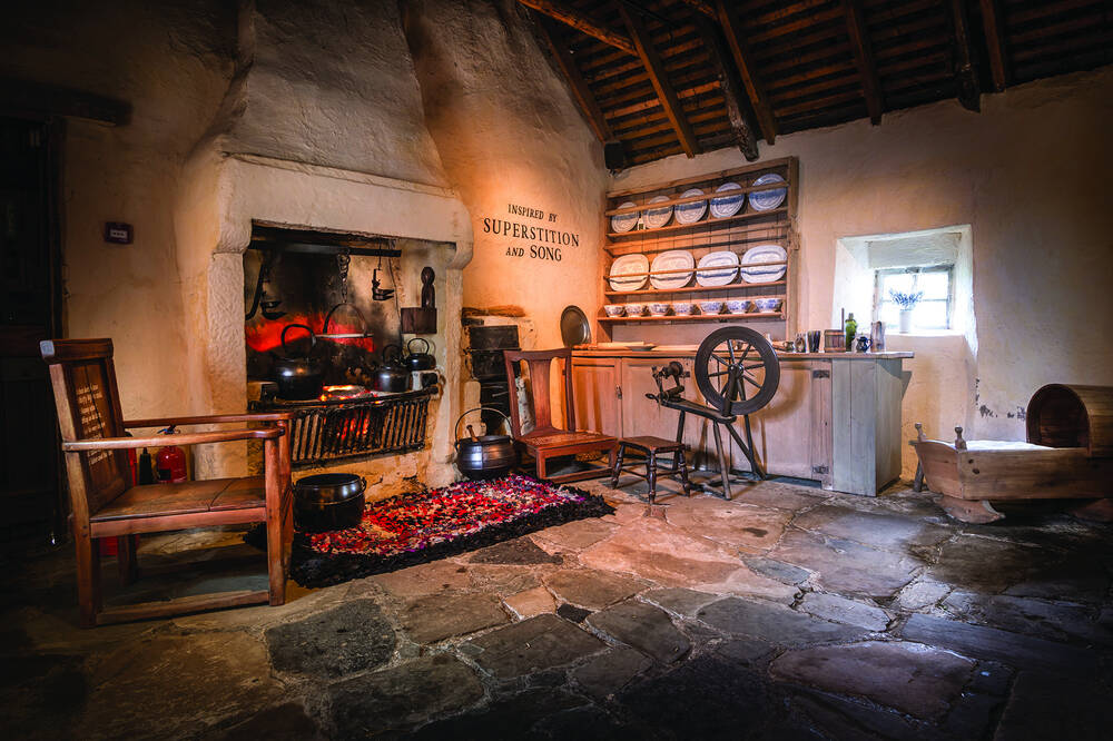 The kitchen at Burns Cottage, with a stone-flagged floor, large stone fireplace and hearth, wooden chairs, and a spinning wheel.