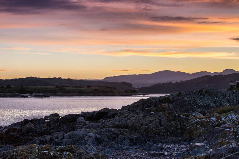 Low tide at sunset, with the rocky shore at Rockcliffe in the foreground. Silhouetted hills can be seen in the distance.