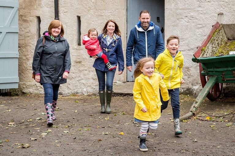 A family walk across a farmyard towards the camera. Two young children in bright yellow coats run ahead.