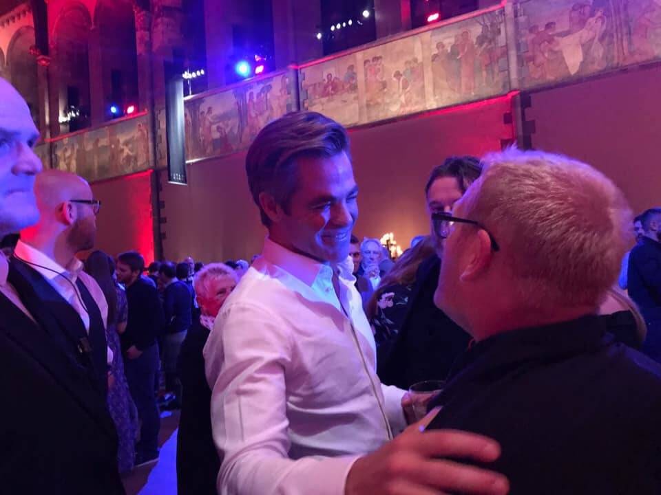 The actor Chris Pine greets another man and claps his hand on his shoulder at an awards ceremony, lit with pink and blue lights.