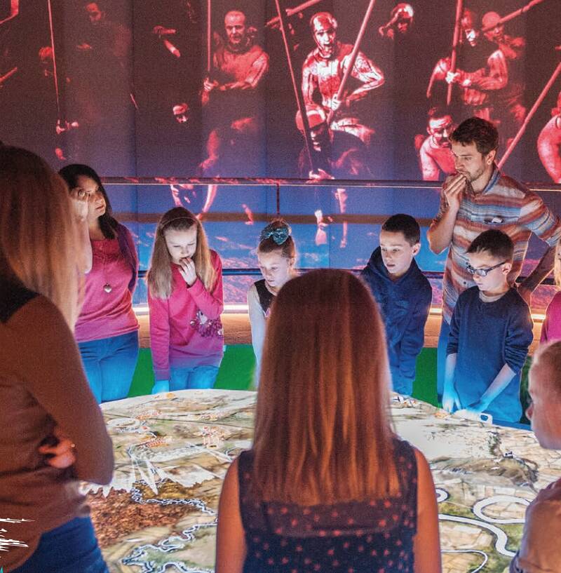 A group of children, with a few adults, stand around the Battle table at Bannockburn Visitor Centre. Images of soldiers are displayed on the wall.