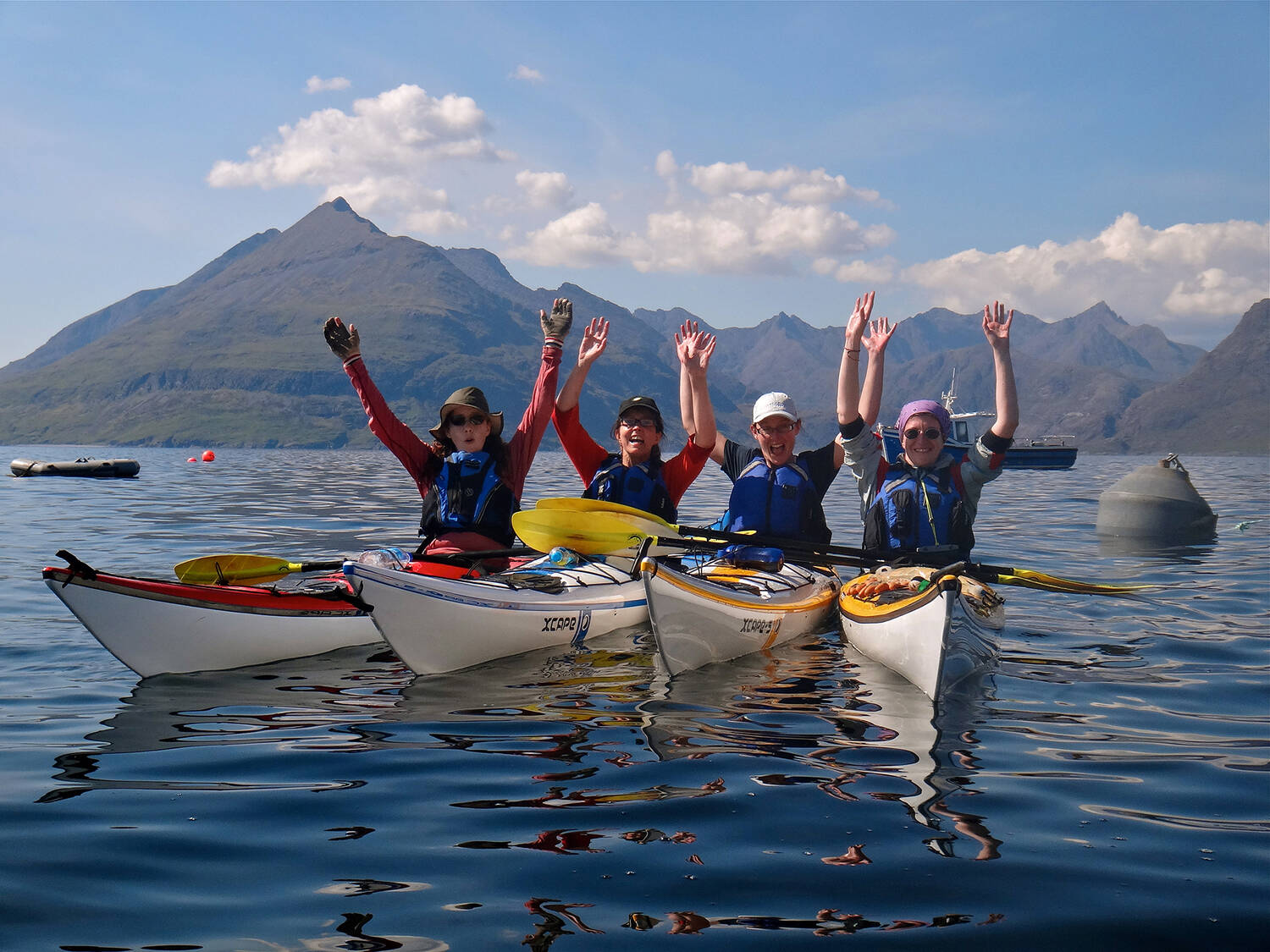 Four people in a row in kayaks raise their hands in the air. The water is very still and blue. Tall mountains rise behind them in the distance.