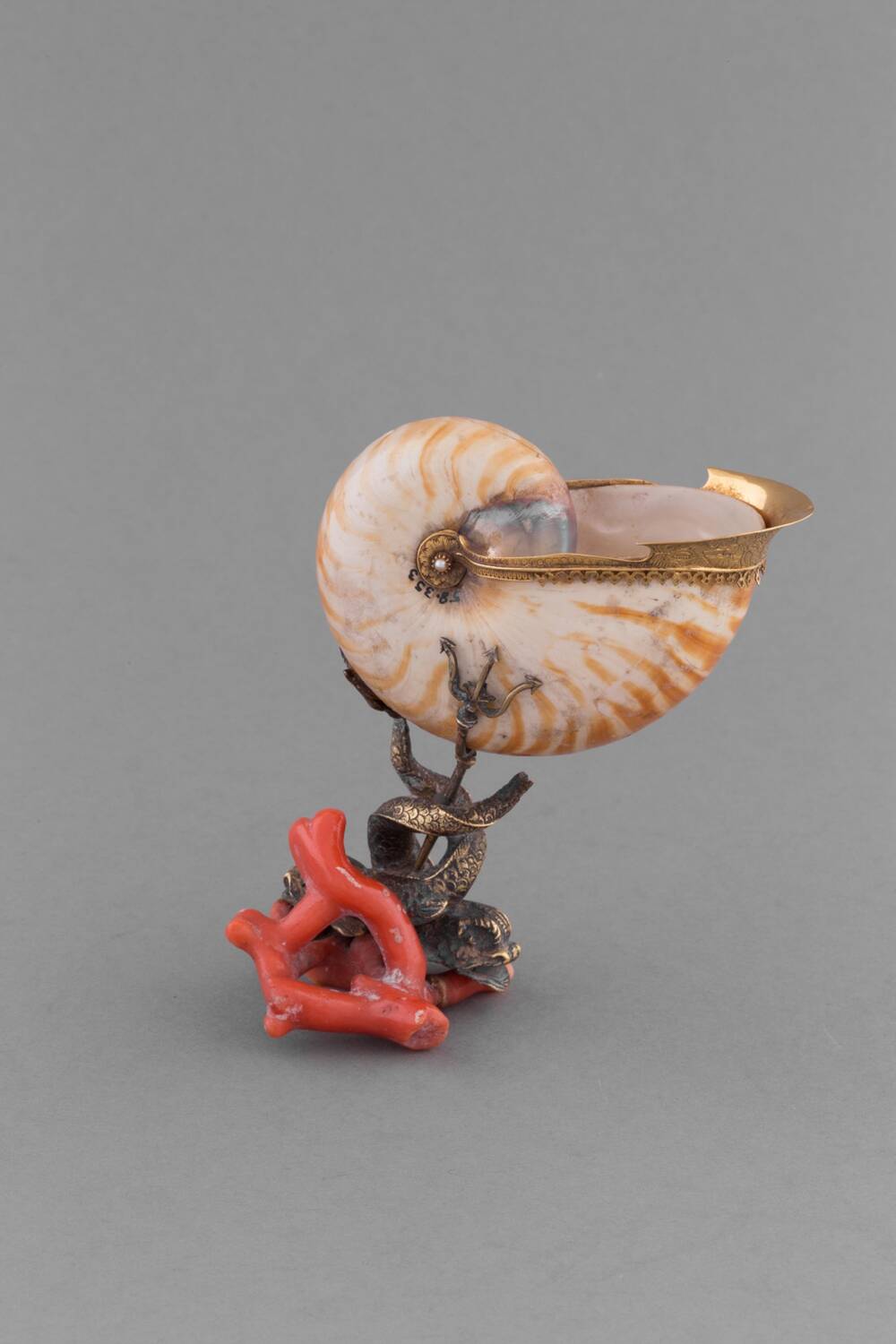 A type of cup made from shells and silver is displayed against a plain grey background. The main body of the cup is made from a large brown and white shell, where the lip has been coated in gold. It is supported by a silver stand, shaped like dolphins and tridents. The base appears to be made of coral.