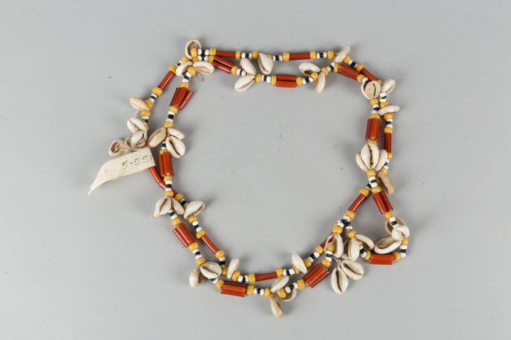 A shell and bead necklace is displayed in a circle against a plain grey background. The shells are small white cowrie shells; the beads are brown, orange or yellow.