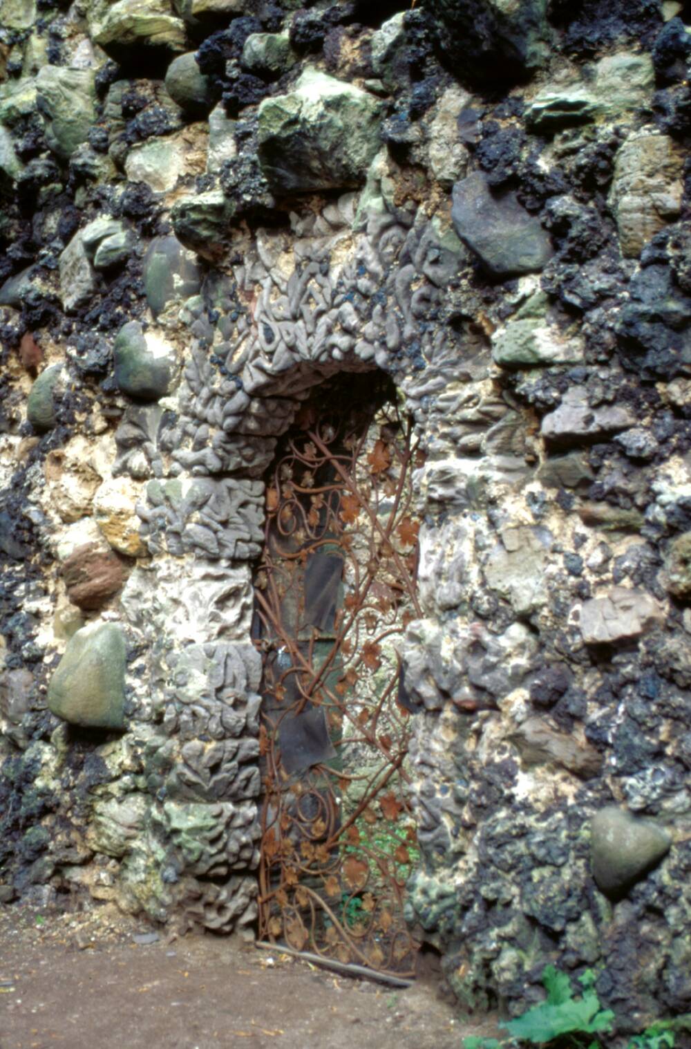 A close-up of the stone doorway to a stone grotto, with many stones and shells stuck to the walls. The gate is made of delicate metal, with leaf carvings throughout.